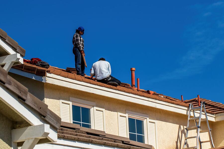 Two workers on the edge of a barrel tile roof under a clear sky