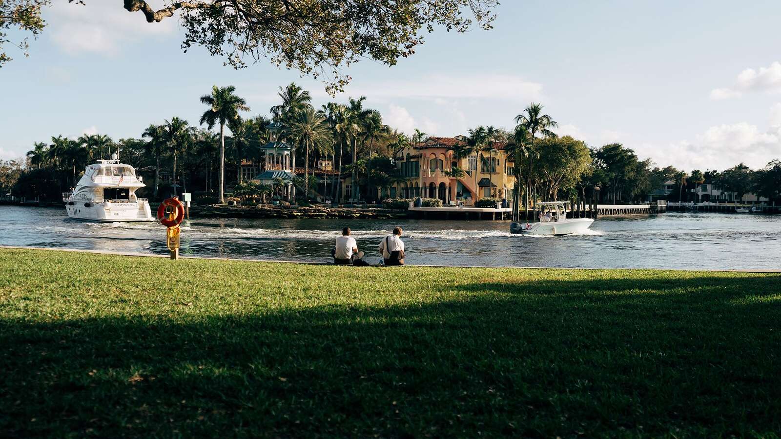 A riverside scene with palms, moored boats and people on the bank